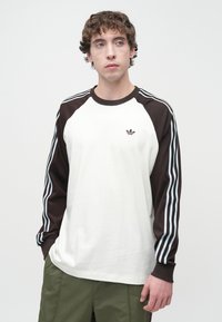 Μπλούζα μπέιζμπολ μακρυμάνικη, λευκή και καφέ, με αντιθετικά raglan μανίκια, που διαθέτει μπλε ρίγες και ένα μικρό καφέ λογότυπο στο στήθος.