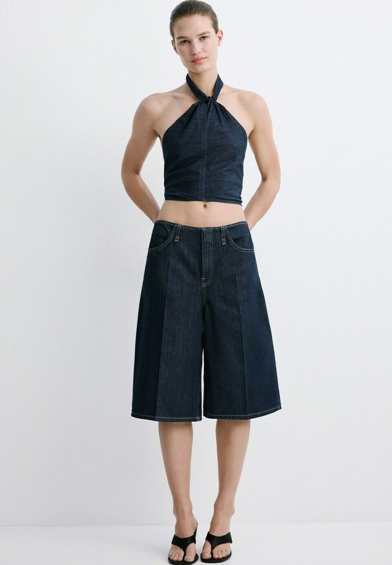 Femme portant un haut sans manches en denim foncé à col halter et un short en denim large jusqu'aux genoux, assortis à des talons ouverts noirs, sur fond blanc.