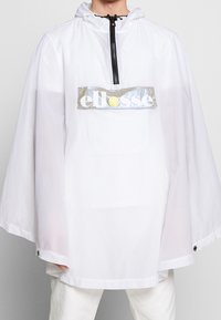 Vit, lätt poncho med huva, svart dragkedjestängning och en reflekterande grå logoplåster. Har tryckknappar i fållen.