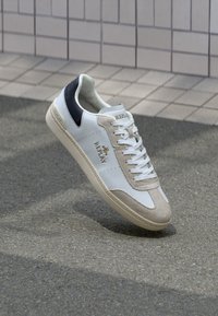 Sneakers bianche a taglio basso con dettagli in suede beige chiaro e un contrafforte blu scuro. Presentano una linguetta testurizzata e un logo sul lato.
