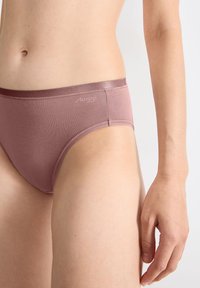 Vrouw in mauve katoen ondergoed, die haar onderlichaam en rechterhand laat zien tegen een effen lichte achtergrond.