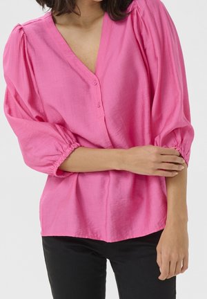 Femme ajustant la manche d'un chemisier ample à col en V de couleur rose vif, associé à un pantalon noir, sur un fond clair uni.