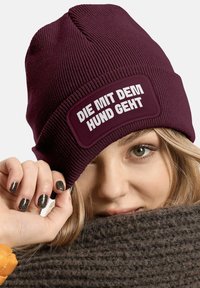 Burgundy knit beanie with a rectangular patch featuring white text, “DIE MIT DEM HUND GEHT,” textured fabric, fitted shape.