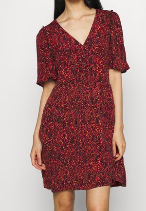 Kobieta ubrana w sukienkę z krótkim rękawem i dekoltem w serek, ozdobioną wzorem czerwonych, pomarańczowych i różowych liści na ciemnym tle.