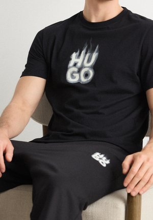 Mann sitzt auf einem Stuhl, trägt ein schwarzes T-Shirt und eine Hose mit weißem "HUGO"-Flammenlogo auf Brust und Oberschenkel.