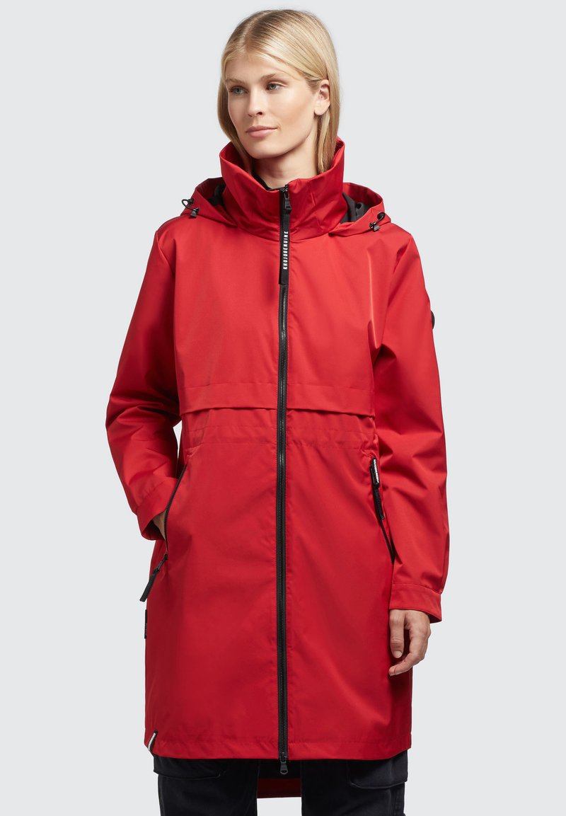 Chaqueta impermeable roja con un alto cuello, capucha ajustable, cremallera frontal y bolsillos laterales. Presenta una textura suave y un corte holgado.