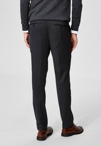 Mørkegrå formelle bukser laget av stoff med en subtil tekstur. Har to baklommer med knappelukking. Kombinert med brune sko.