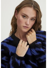 Črno-modra prugasta pulover s mehko, puhaste teksturo. Ima globok V-izrez in široke manšete, tesno prilega ramenom.