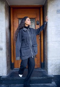 Kobieta z długimi włosami w za dużej ciemnej kurtce, czarnych spodniach i białych butach schodząca po kamiennych schodach obok drewnianych drzwi, ręka oparta na ścianie.