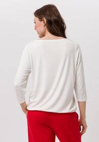 Femme aux longs cheveux bruns portant un haut à manches longues blanc et un pantalon rouge, se tenant avec les mains près des poches, dos à l'appareil photo.