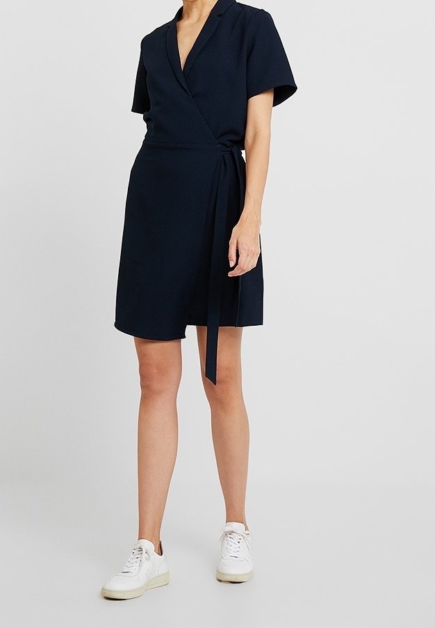 Mujer vestida con un vestido envolvente de manga corta en azul marino, con un lazo en la cintura y zapatillas blancas, de pie frente a un fondo blanco liso.