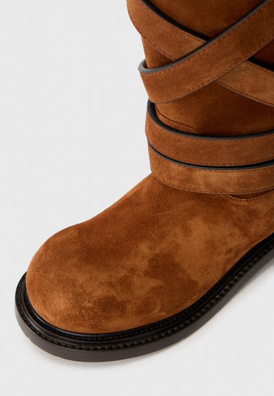 Brown suede ankle boot with black sole and decorative crisscross straps above the ankle.