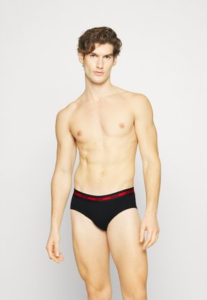 Slip neri con fascia elastica rossa che presenta il logo "HUGO". Tessuto liscio, design aderente e taglio basso. Il modello mostra la vista frontale.