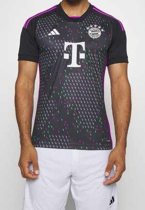 Mann, der ein schwarzes FC Bayern München Fußballtrikot mit lila-grünem Muster, weißem Sponsorlogo und weißen Shorts trägt.