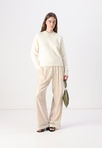 Maglione crema, taglio morbido e ampio, texture soffice; pantaloni beige con paillettes lucide; borsa verde dalla forma arcuata; sandali neri.
