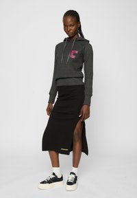 Sudadera gris corta con "ESPERANDO EL VIERNES" en texto rosa, combinada con una falda midi negra con una abertura lateral y el logo de la marca en amarillo. Zapatillas negras.