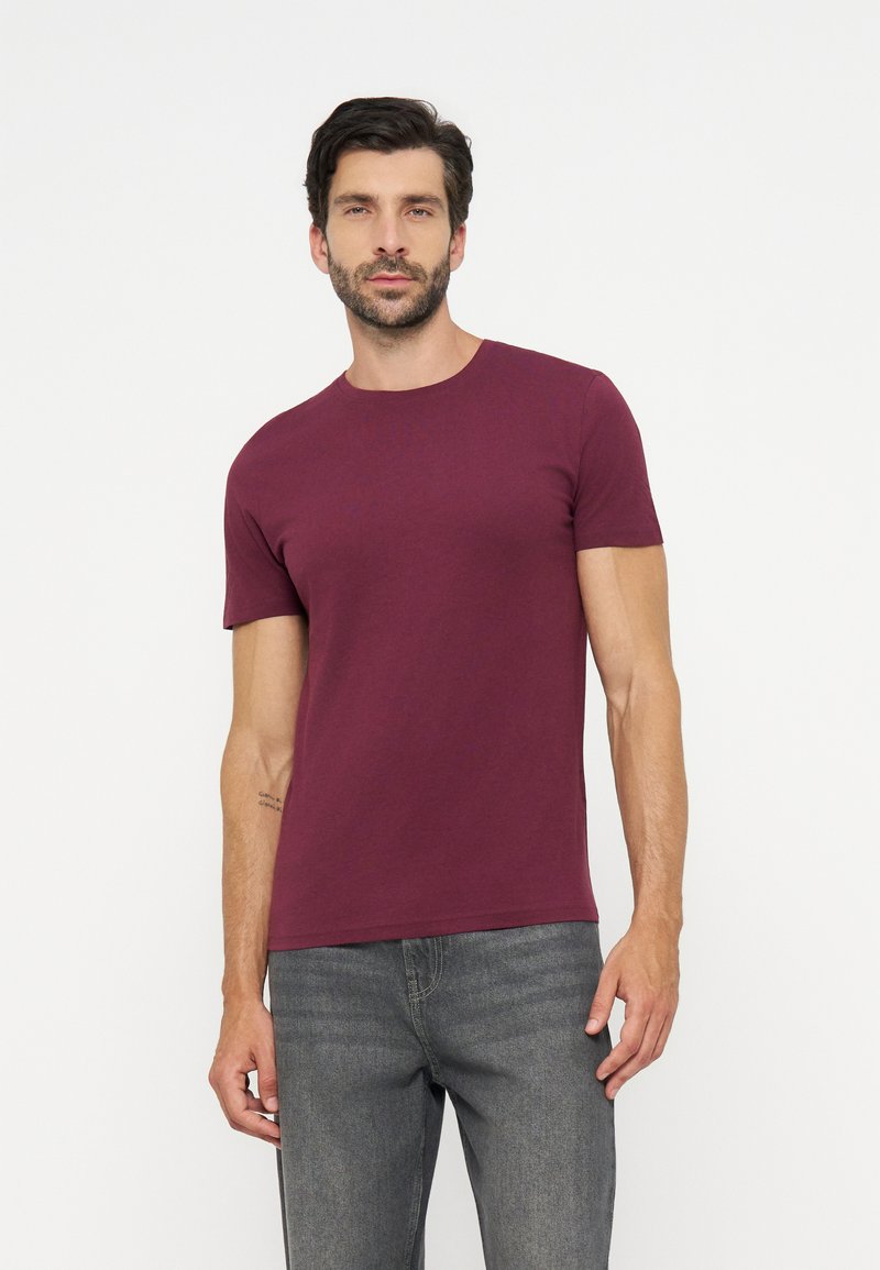 Man met donker haar en baard, die een effen bordeauxrood T-shirt met korte mouwen en grijze spijkerbroek draagt, staand tegen een witte achtergrond.