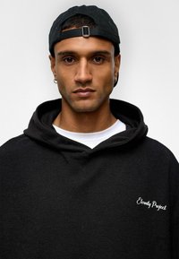 Sweatshirt à capuche noir avec un tissu texturé, présentant un logo brodé blanc "Eternity Project" sur le devant. Superposé à une chemise blanche.