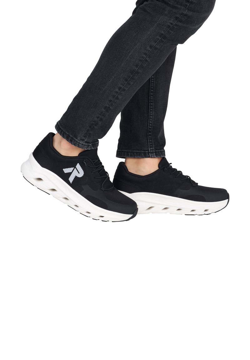 Personne portant des chaussures de sport noires avec des semelles blanches et un jean noir sur fond blanc.