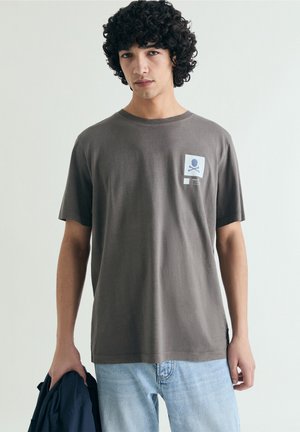 Jeune homme aux cheveux bouclés portant un T-shirt gris imprimé tête de mort et os croisés, et un jean bleu clair, tenant une veste sombre dans une main.