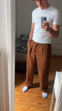 Jovem vestido com uma t-shirt branca, calças castanhas largas e meias brancas, tirando uma selfie no espelho num quarto com chão de madeira.