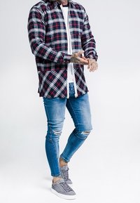 Hombre con camisa de cuadros rojo y azul marino, camiseta blanca, jeans ajustados rasgados, zapatillas grises, ajustándose las manos tatuadas.