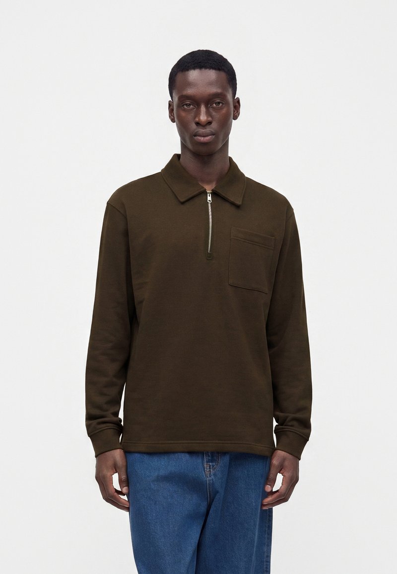 Homme portant un polo à manches longues marron foncé avec une fermeture éclair à l'avant et une poche, associé à un jean en denim bleu, debout devant un fond uni.