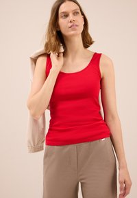 Canottiera rossa con scollatura a barca, realizzata in tessuto liscio. Abbinata a pantaloni beige texture, con una giacca beige casual appoggiata su una spalla.