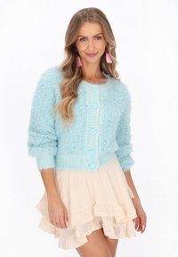 Kobieta z falującymi blond włosami nosi różowe kolczyki z frędzlami, puszysty jasnoniebieski sweter zapinany na guziki oraz beżową spódnicę warstwową z falbanami.