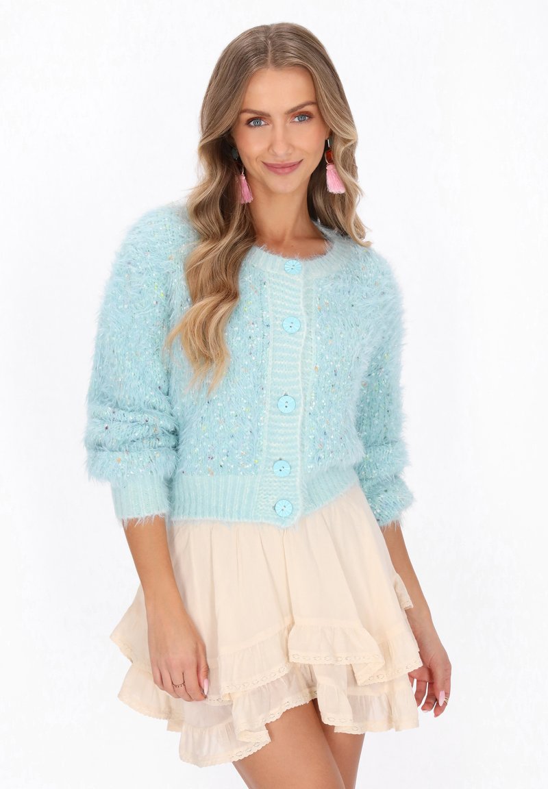 Kobieta z falującymi blond włosami nosi różowe kolczyki z frędzlami, puszysty jasnoniebieski sweter zapinany na guziki oraz beżową spódnicę warstwową z falbanami.