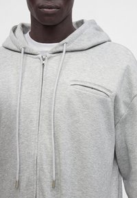 Personne à la peau foncée portant un sweat à capuche gris clair zippé avec cordons et une petite poche poitrine sur le côté gauche.