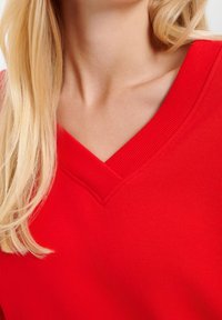 Blonde vrouw in een felrode V-hals trui, met een blootgestelde nek en schoudergebied, met los haar aan één kant.