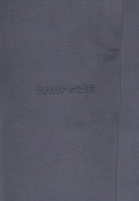 Tissu gris foncé avec logo texte en relief « DROPSIZE » cousu près d’une couture verticale.