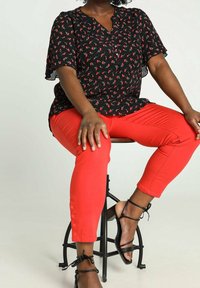 Personne assise sur un tabouret, portant un pantalon rouge court avec des boutons, un chemisier noir à motifs floraux et des sandales noires à brides.