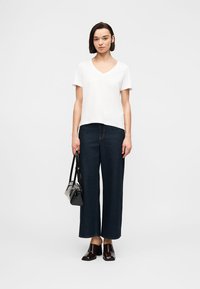 Γυναίκα που φοράει λευκό T-shirt με V-λαιμόκοψη, σκούρα φαρδιά τζιν, μαύρα παπούτσια με τακούνι, κρατώντας μια μαύρη τσάντα, μπροστά σε απλό φόντο.