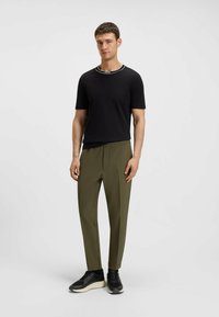 Schwarzes T-Shirt mit kurzen Ärmeln und weißen Kragenakzenten, kombiniert mit olivgrünen, schmal zulaufenden Hosen und schwarzen Schuhen mit dicker Sohle.