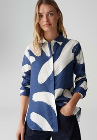 Blau-weiß gemustertes Hemd mit Kragen, das markante Pinselstrich-Designs aufweist. Hergestellt aus glattem Stoff, mit Knopfdetails und hochgekrempelten Ärmeln.