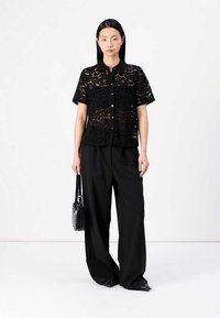 Chemisier noir en dentelle à manches courtes avec un bouton devant, porté avec un pantalon large noir. Le modèle tient un petit sac à main noir.