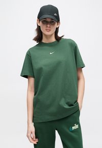 Νέος άνθρωπος φορώντας μπλούζα Nike σκούρου πράσινου χρώματος και παντελόνι με λογότυπα, και μαύρο καπέλο Nike, στέκεται με το ένα χέρι στην τσέπη.