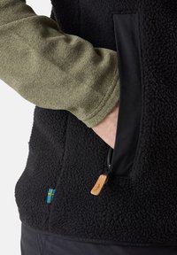 Svart fleecejacka med en texturerad yta, dragkedjefickor på sidorna med läderdragkedjor och en grön långärmad tröja under.
