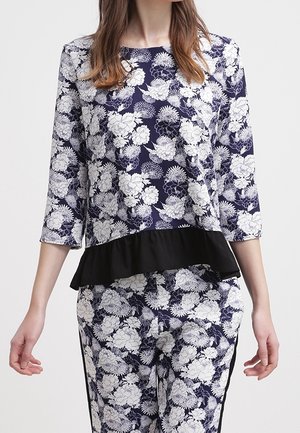 Femme portant un haut fleuri bleu marine et blanc avec manches trois-quarts et un pantalon assorti, avec un détail volant noir à la taille.