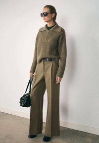 Suéter de punto acanalado color oliva con cuello con cremallera, pantalones amplios color beige con pliegues, bolso negro y gafas de sol. Textura suave en todo.