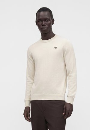 Homme portant un pull crème à manches longues avec un petit logo zèbre sur la poitrine et un pantalon marron foncé sur un fond clair uni.