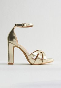 Metallisk gyllene högklackad sandal med ankelrem och korslagd frontdesign. Har en kraftig klack och en mjuk beige innersula.