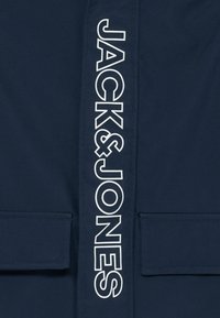 Veste bleue marine à l'avant avec le texte vertical blanc "JACK & JONES" et deux poches à rabat près du bas.