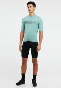 Maillot de cyclisme turquoise avec une fermeture éclair partielle et des détails de rayures horizontales, associé à un short rembourré noir et des chaussures de cyclisme.