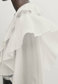 Blusa bianca con strati di rouches lungo le maniche, realizzata in un tessuto leggero e trasparente che cade morbido. Design minimalista.