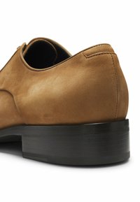 Brauner Wildlederschuh mit Absatz und schwarzer gestapelter Sohle, sichtbare Naht, dargestellt vor weißem Hintergrund.