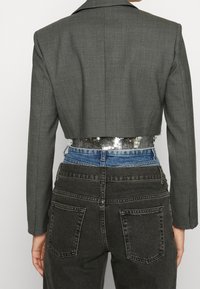 Blazer gris ajustado con un diseño estructurado, que presenta una cinturilla acentuada con lentejuelas, combinado con jeans oscuros de talle alto.