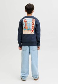 Persona in piedi di spalle che indossa una felpa blu scuro con una grafica colorata di un tempio di Bangkok e jeans blu chiaro a gamba larga.